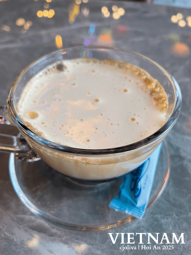 Glass cup of Vietnamese egg coffee in Hoi An, filled with creamy froth, placed on a glass saucer with a blue packet, against a marble surface and colorful bokeh lights.