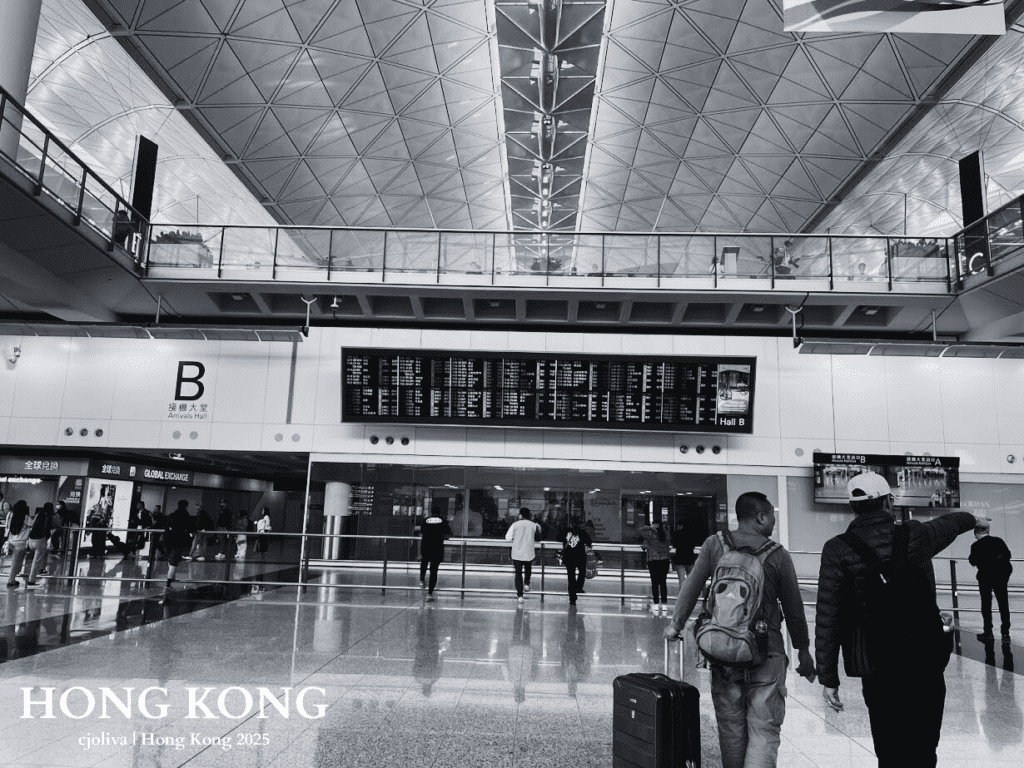 Black and white photo of Hong Kong International Airport Arrivals Hall B with geometric ceiling, flight information display, travelers with luggage, and shops like Global Exchange.