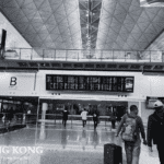 Black and white photo of Hong Kong International Airport Arrivals Hall B with geometric ceiling, flight information display, travelers with luggage, and shops like Global Exchange.