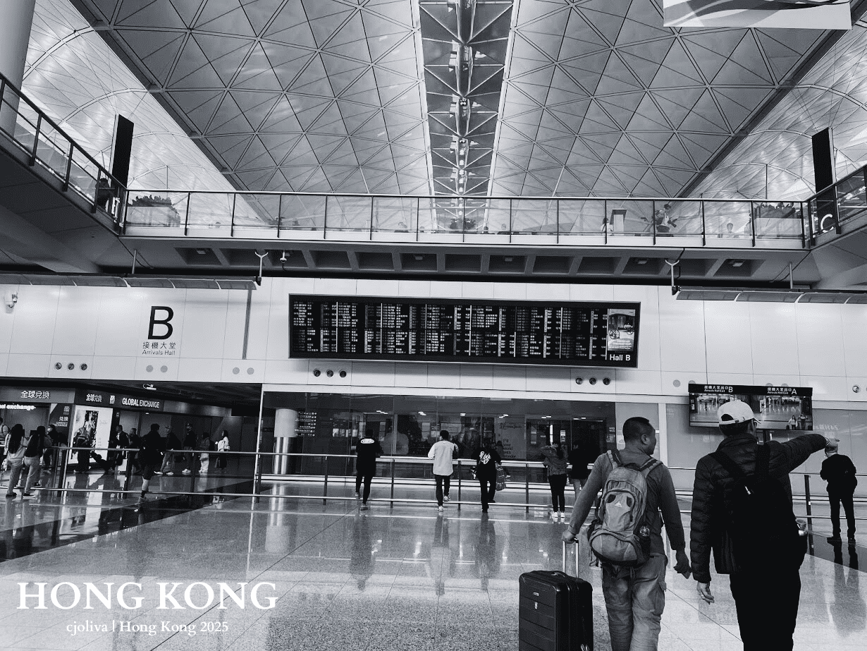 Black and white photo of Hong Kong International Airport Arrivals Hall B with geometric ceiling, flight information display, travelers with luggage, and shops like Global Exchange.
