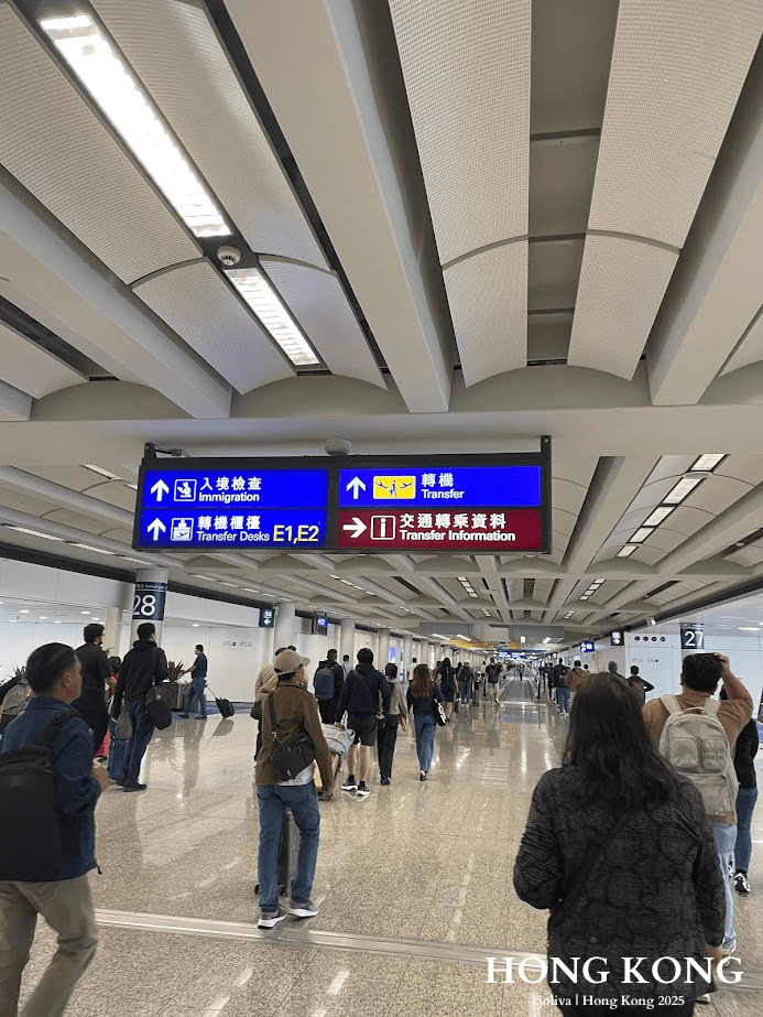 Busy corridor at Hong Kong International Airport with travelers walking under bilingual signs directing to immigration, transfer desks, and gates.
