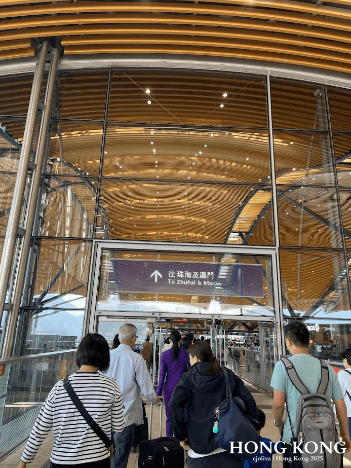 Travelers walking into a modern Hong Kong border terminal with a curved wooden ceiling and signage reading "To Zhuhai & Macao" in English and Chinese.