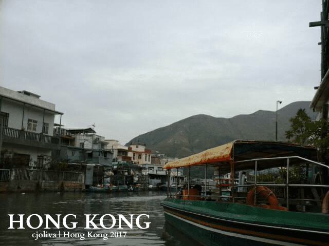 A green and yellow boat with a canopy docked on a canal in Hong Kong, surrounded by multi‑story residential buildings and green hills under a cloudy sky.