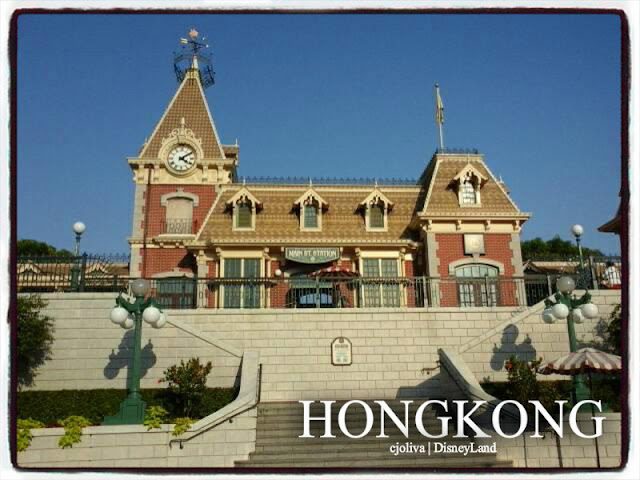 Victorian-style Main Street Train Station at Hong Kong Disneyland with a clock tower, gabled roofs, a staircase, and lamp posts under a clear blue sky.