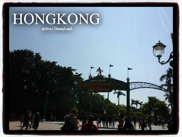 Entrance arch to Hong Kong Disneyland Resort with Mickey Mouse figure, flags, palm trees, and visitors walking toward the park.