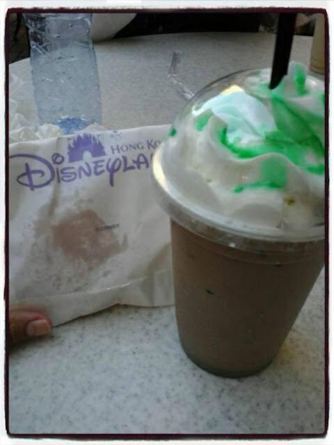 A chocolate or coffee drink with whipped cream and green syrup in a clear cup beside a Hong Kong Disneyland wrapper with a castle logo.