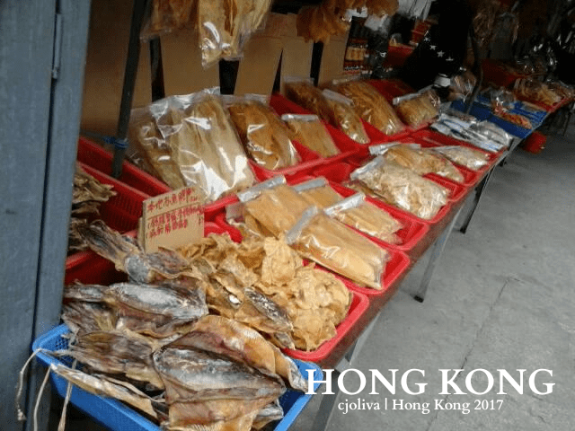Market stall in Hong Kong selling dried seafood, including fish, squid, and preserved marine products in red baskets and clear plastic bags.