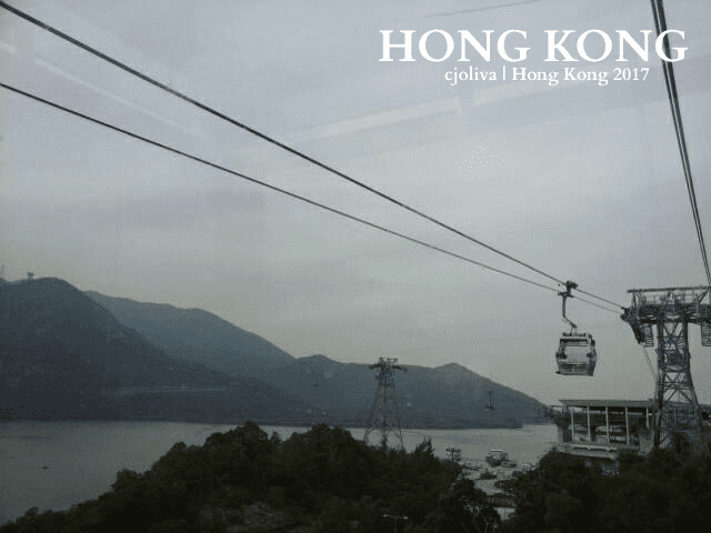 Ngong Ping 360 cable cars suspended over water and lush mountains in Hong Kong, with misty peaks and cloudy sky.
