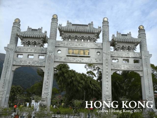 Traditional Chinese stone archway with ornate carvings and roofs at the entrance to Po Lin Monastery in Hong Kong, surrounded by trees and mountains.