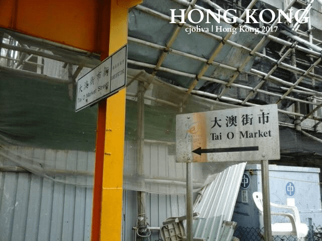 Street signs in Hong Kong directing visitors to Tai O Market and Tai O Market Street, written in Chinese and English, with scaffolding and construction in the background.