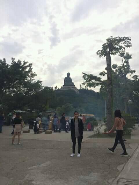 Large seated Tian Tan Buddha statue on Lantau Island, Hong Kong, surrounded by trees with visitors in the foreground under a cloudy sky.