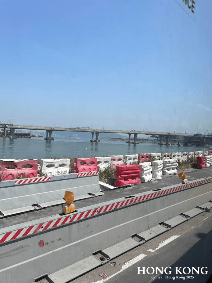 Hong Kong waterfront construction zone with Gammon barriers, warning lights, and a multi-span bridge extending across calm water under a clear blue sky.