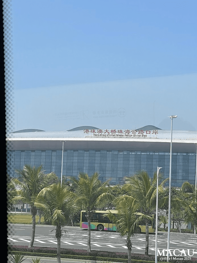 Exterior view of the Hong Kong–Zhuhai–Macau Bridge Zhuhai Port with a modern curved roof, glass facade, palm trees, and colorful bus under a clear blue sky.