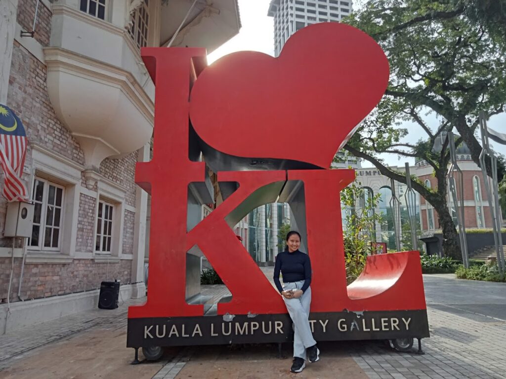 I Love KL red sculpture at Kuala Lumpur City Gallery, with the colonial-style building, Malaysian flag, and modern skyscrapers in the background.