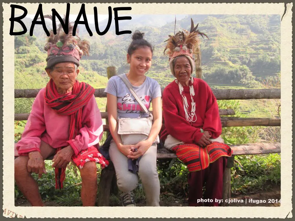 A traveler sitting on a wooden bench between two elderly Ifugao women wearing traditional feathered headdresses and red woven clothing, with rice terraces in the background.