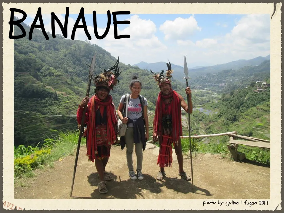 A traveler posing between two Ifugao men in traditional red woven attire and feathered headdresses, holding spears, with the Banaue Rice Terraces in the background.