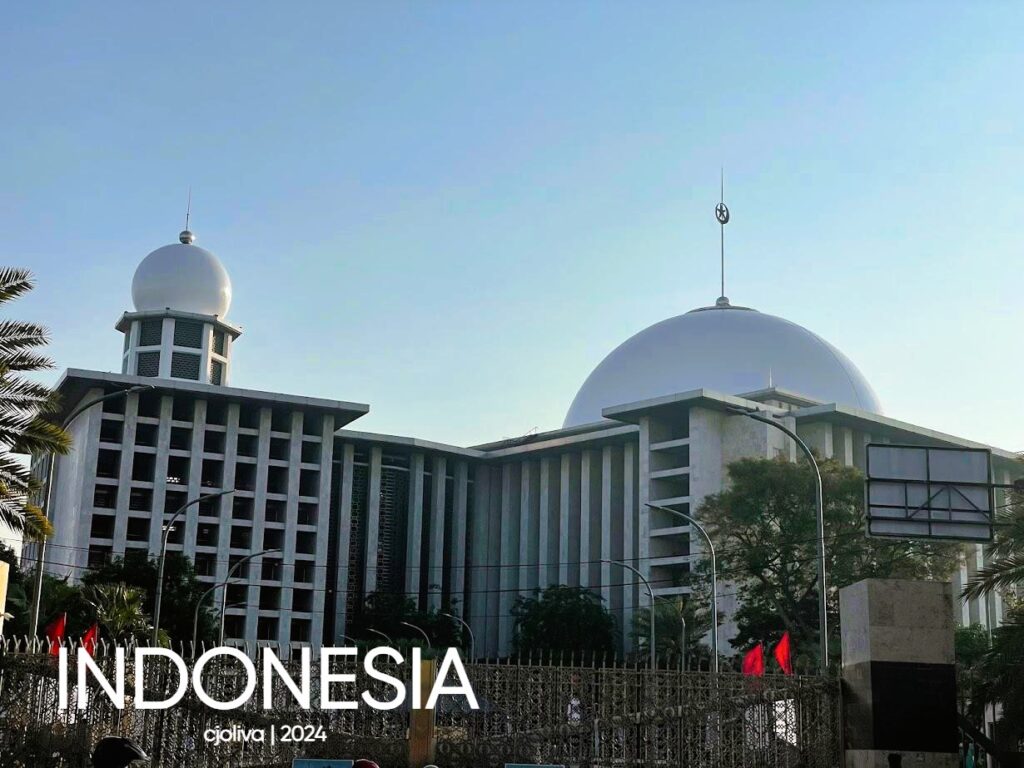 The Istiqlal Mosque in Jakarta, showing its large white main dome and a smaller domed minaret. The building features a modern, rectangular architectural style with vertical pillars and is surrounded by palm trees.