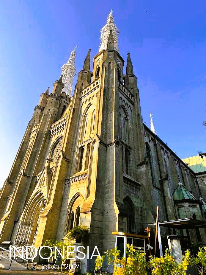 The stone exterior and tall white metal spires of the Jakarta Cathedral, a neo-Gothic Catholic church, viewed from a low angle with yellow flowers in the foreground and a clear blue sky.