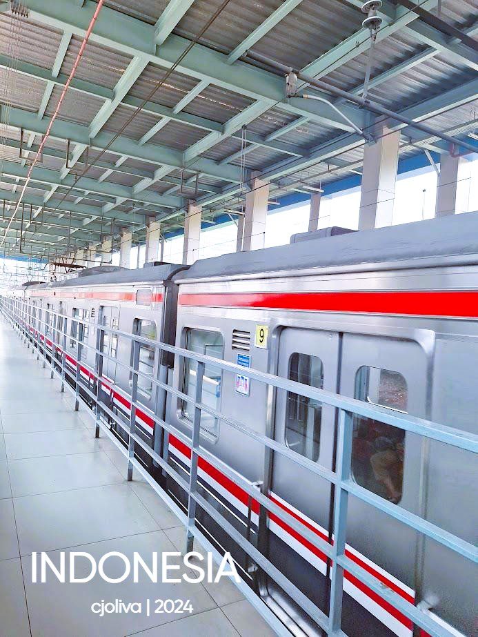 Silver commuter train with red and white stripes at a modern Indonesian station platform, covered by a metal roof with beams and columns.