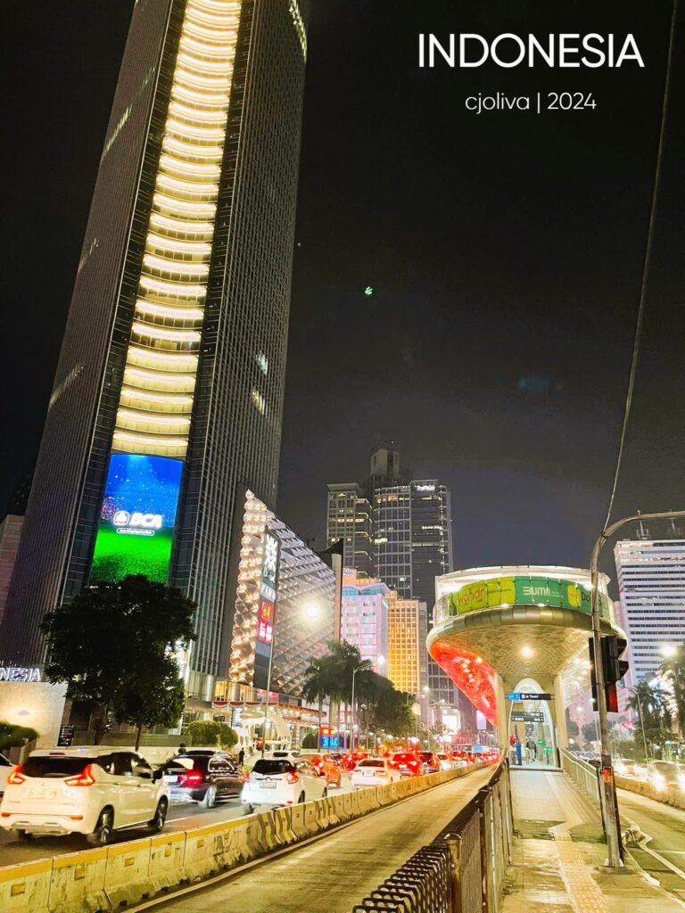 A vertical night photography shot of a busy street in Jakarta, Indonesia, featuring a tall skyscraper with a blue LED screen, glowing city traffic, and a modern, rounded pedestrian bridge/bus station illuminated in gold and green.