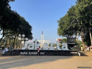 A large white 3D sign reading "Jakarta kota kolaborasi" with the West Irian Liberation Monument in the background under a clear blue sky.