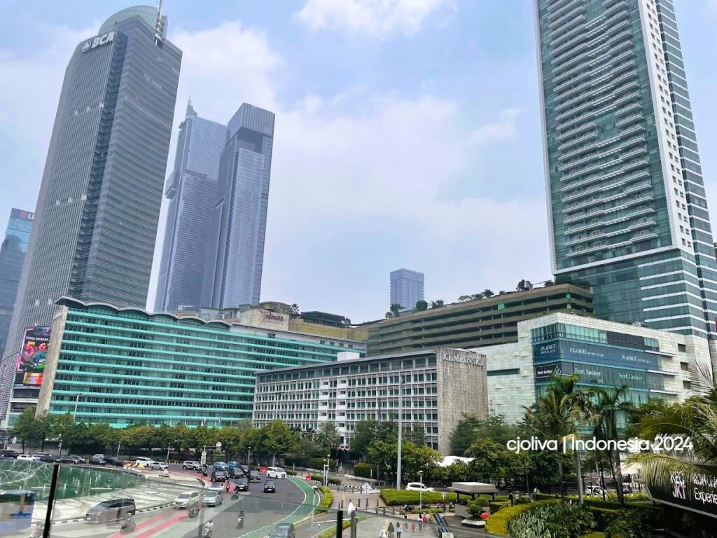 A high-angle view of modern glass skyscrapers, including Menara BCA and Hotel Indonesia Kempinski, surrounding the Bundaran HI area in Jakarta.