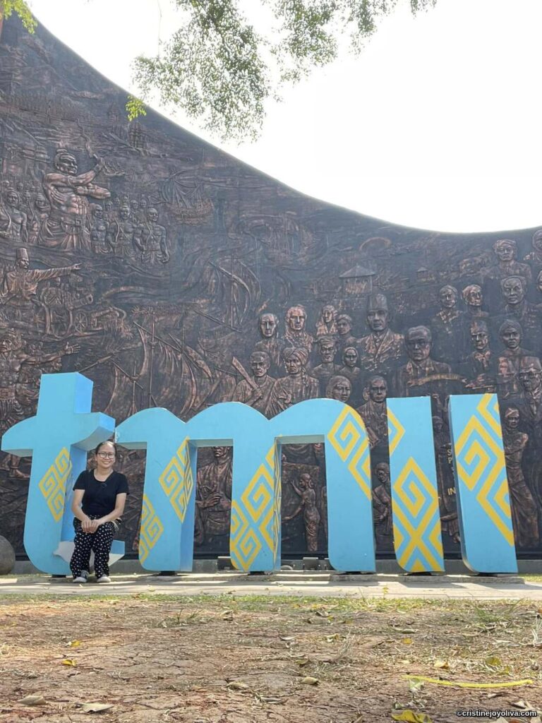 A person sitting in front of large “tmii” letters with yellow geometric patterns and a bronze relief wall depicting Indonesian history at Taman Mini Indonesia Indah, Jakarta.