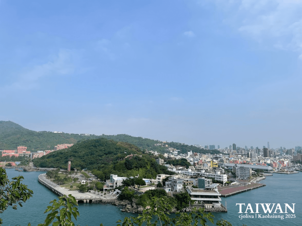 Scenic view of Kaohsiung city with green hill, waterfront promenade, and skyline of tall buildings under clear blue sky in Taiwan