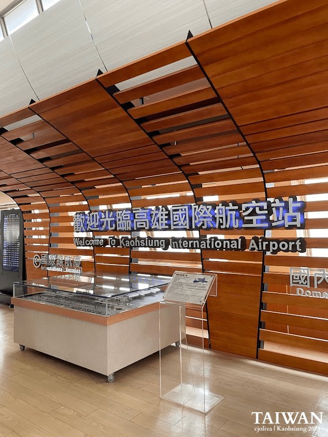 Welcome display at Kaohsiung International Airport with wooden slat wall, bilingual signage, glass diorama, and flight information board