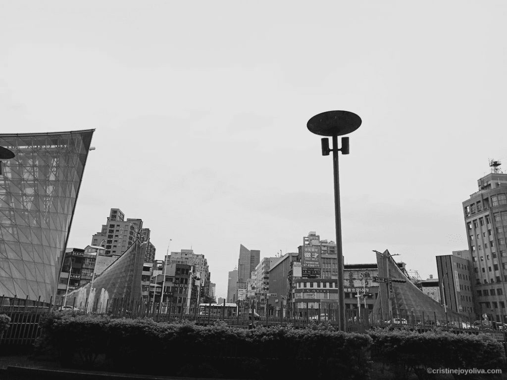 A black and white photograph of the Kaohsiung skyline featuring the modern glass structures of the Formosa Boulevard station entrance under a cloudy sky.