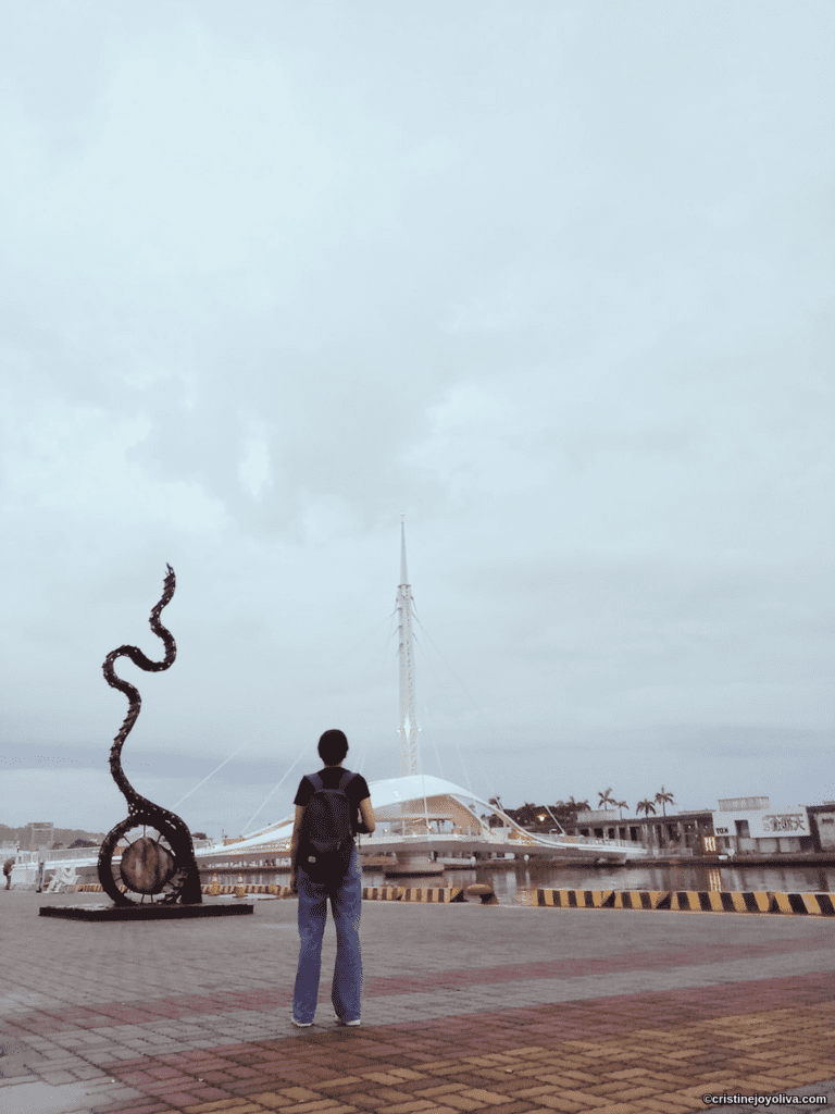 Waterfront view in Kaohsiung, Taiwan with a twisting abstract sculpture, modern white cable‑stayed bridge, palm trees, and buildings under an overcast sky.