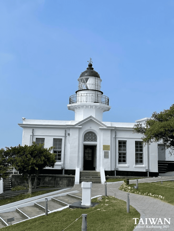White lighthouse with black lantern room, arched entrance, and landscaped walkway under clear blue sky in Kaohsiung, Taiwan