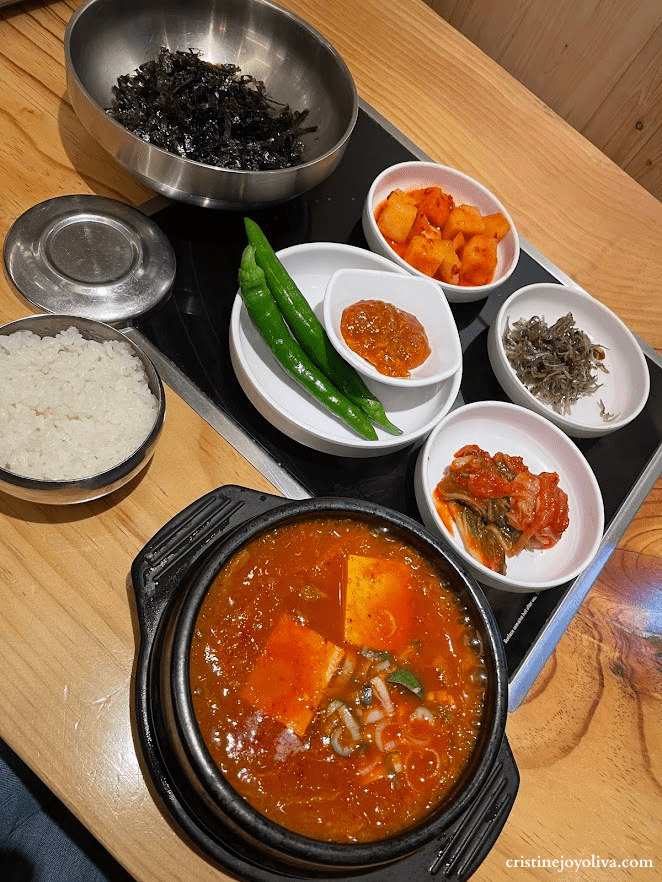 A top-down view of a Korean meal featuring Kimchi Jjigae stew in a black stone pot, a bowl of white rice, shredded seaweed, and five small plates of banchan, including kimchi and green chilies, on a wooden table.