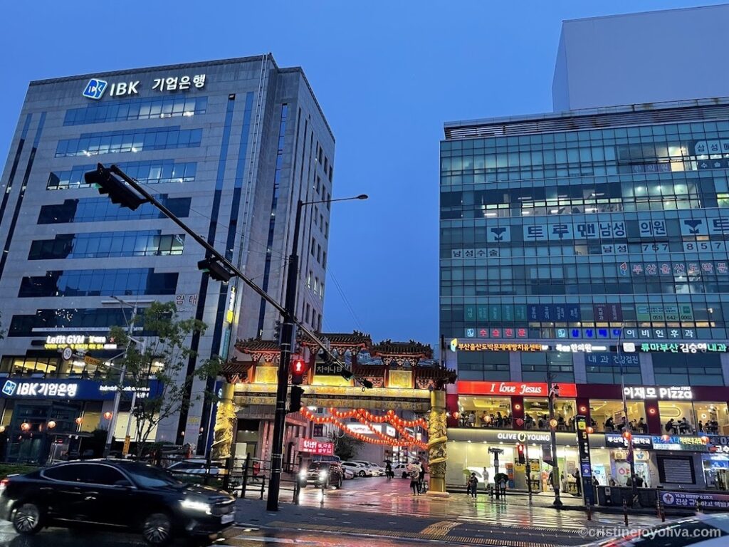 Evening street in Korea with traditional Chinese-style gate, red lanterns, modern buildings, wet pavement, and pedestrians