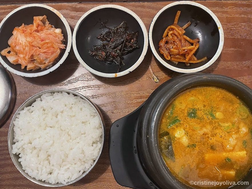 Traditional Korean meal with rice, bubbling stew in stone pot, and side dishes of kimchi, seaweed, and pickled radish