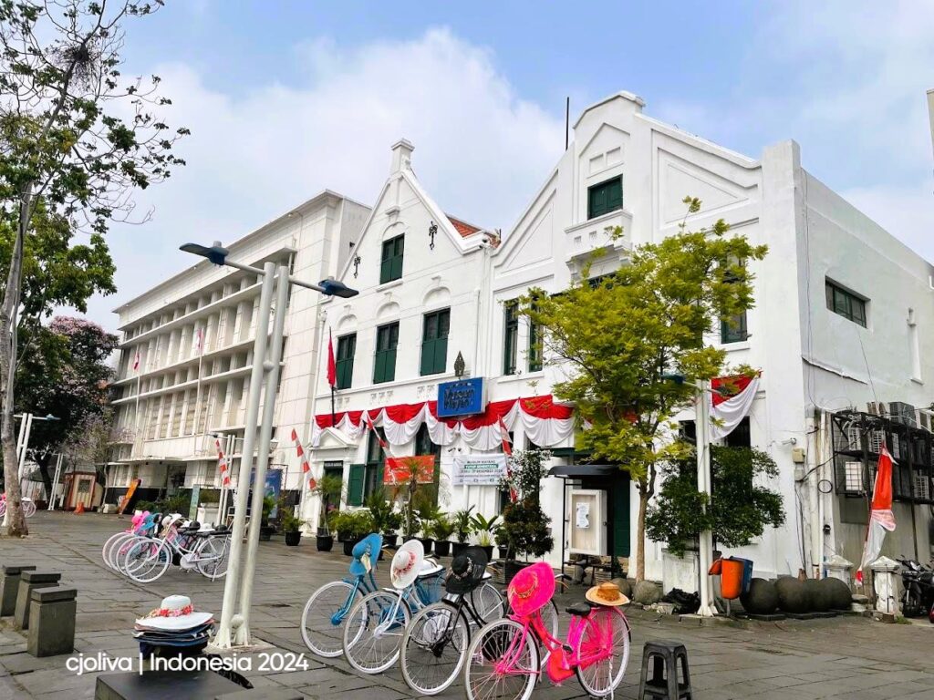 The white colonial-style building of the Wayang Museum in Kota Tua, Jakarta. In the foreground, several colorful vintage bicycles with decorated sun hats are parked on a stone-paved square under a soft sky.