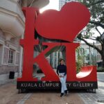 Person sitting in front of the large red “I ❤️ KL” sculpture outside Kuala Lumpur City Gallery, with brick building and trees in the background.