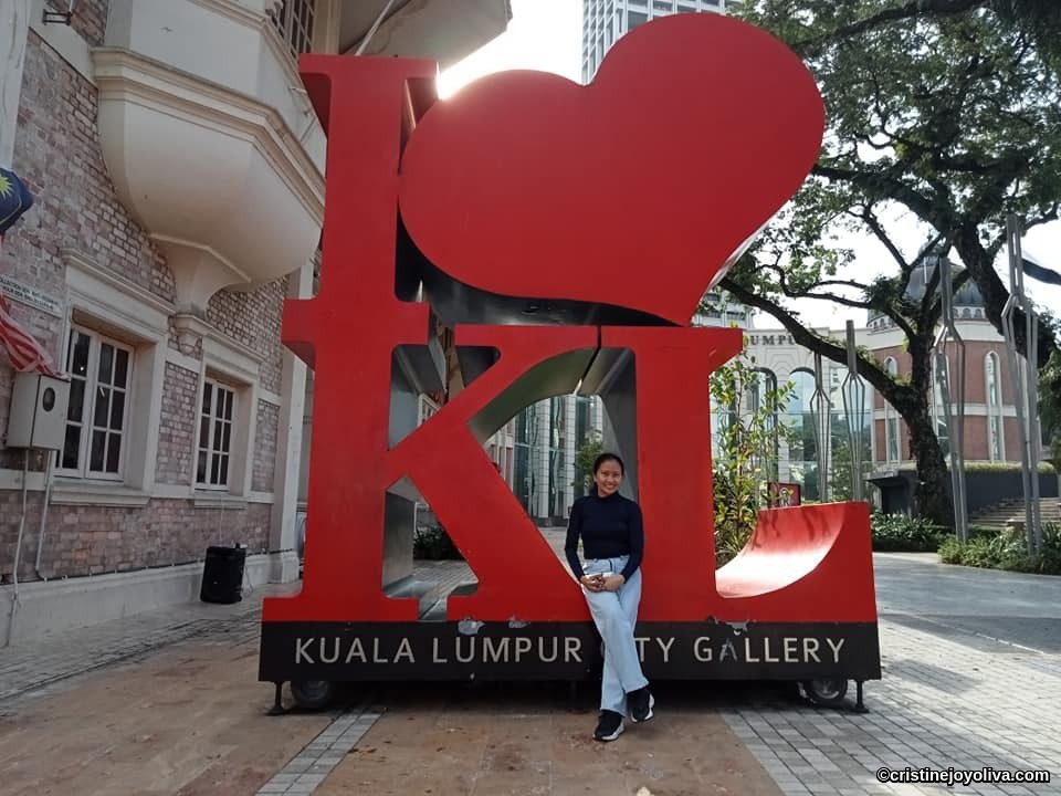 Person sitting in front of the large red “I ❤️ KL” sculpture outside Kuala Lumpur City Gallery, with brick building and trees in the background.