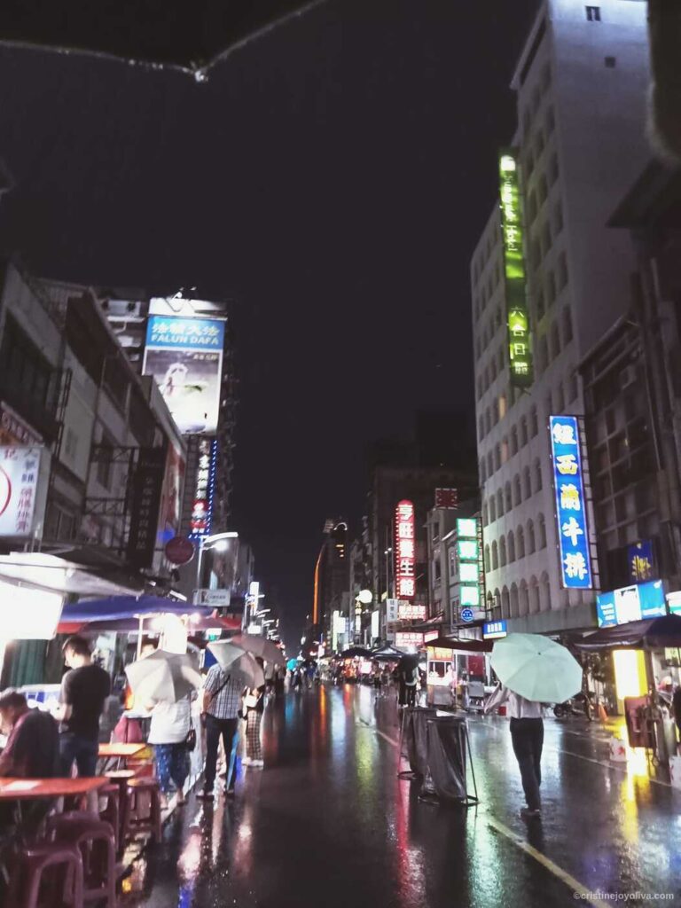 Rainy night at Liuhe Night Market in Kaohsiung, Taiwan, showing wet asphalt reflecting colorful neon shop signs, people with umbrellas, and the Sunnyside Hotel building.