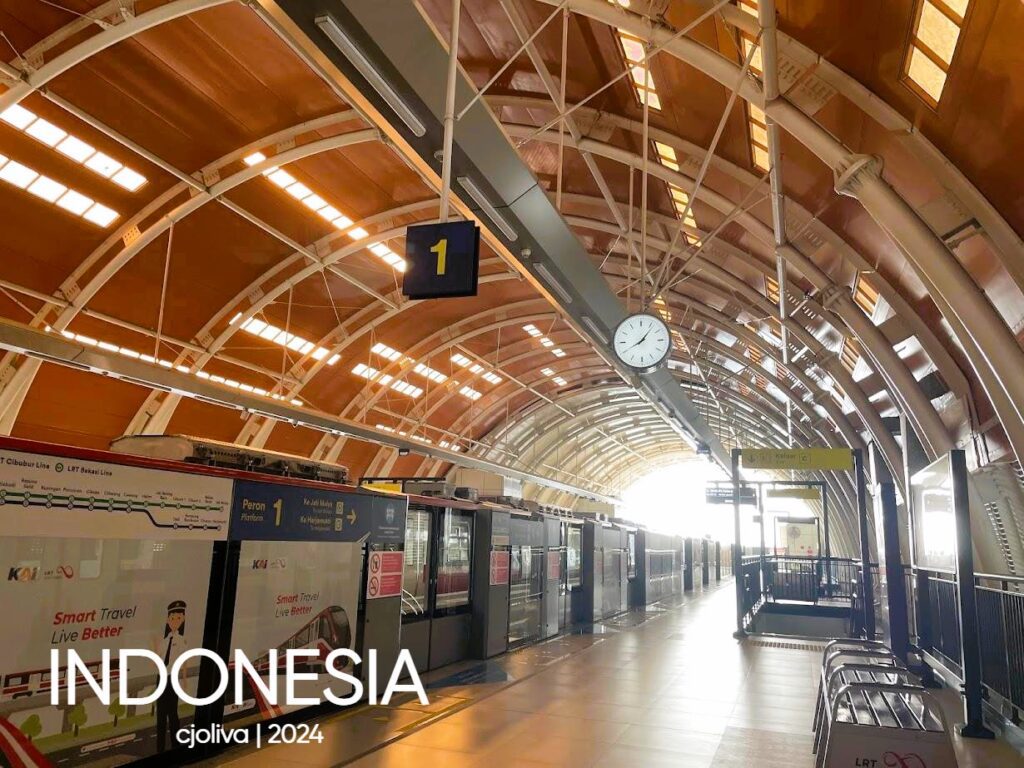 The interior of a high-tech light rail transit (LRT) station in Jakarta. The shot features a high, curved orange ceiling with geometric supports, a clean platform with glass safety doors, and a round clock hanging from the center.