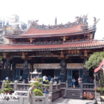 The main hall of Lungshan Temple in Taipei, Taiwan, featuring an ornate orange-tiled roof with dragon sculptures, carved stone columns, and worshippers in the courtyard.