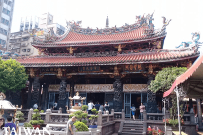 The main hall of Lungshan Temple in Taipei, Taiwan, featuring an ornate orange-tiled roof with dragon sculptures, carved stone columns, and worshippers in the courtyard.