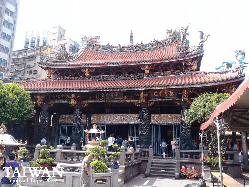 The main hall of Lungshan Temple in Taipei, Taiwan, featuring an ornate orange-tiled roof with dragon sculptures, carved stone columns, and worshippers in the courtyard.