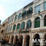 Street in Macau with colonial-style pastel buildings featuring arched windows, green shutters, and retail shops like Bossini and Sasa, with pedestrians and a modern high-rise in the background.