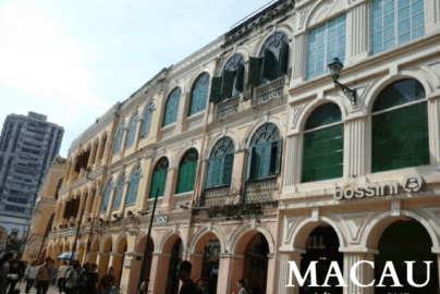 Street in Macau with colonial-style pastel buildings featuring arched windows, green shutters, and retail shops like Bossini and Sasa, with pedestrians and a modern high-rise in the background.