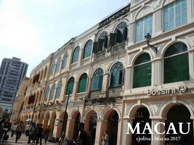 Street in Macau with colonial-style pastel buildings featuring arched windows, green shutters, and retail shops like Bossini and Sasa, with pedestrians and a modern high-rise in the background.
