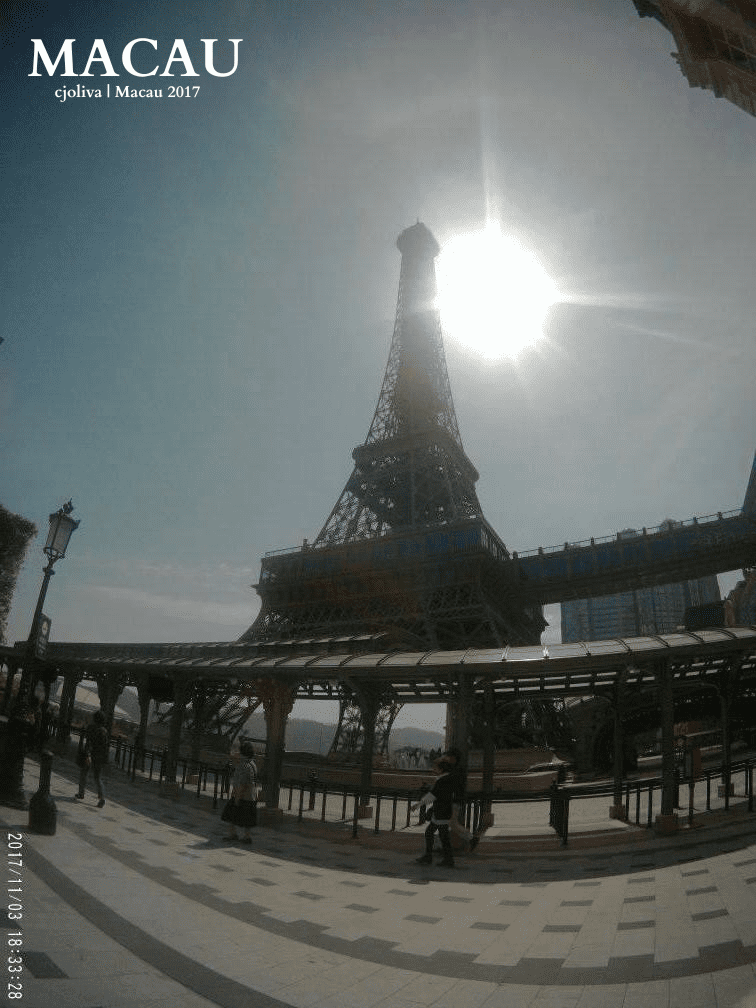Replica of the Eiffel Tower in Macau, China, photographed with the sun backlighting the top of the structure, surrounded by pedestrian walkways and clear skies.