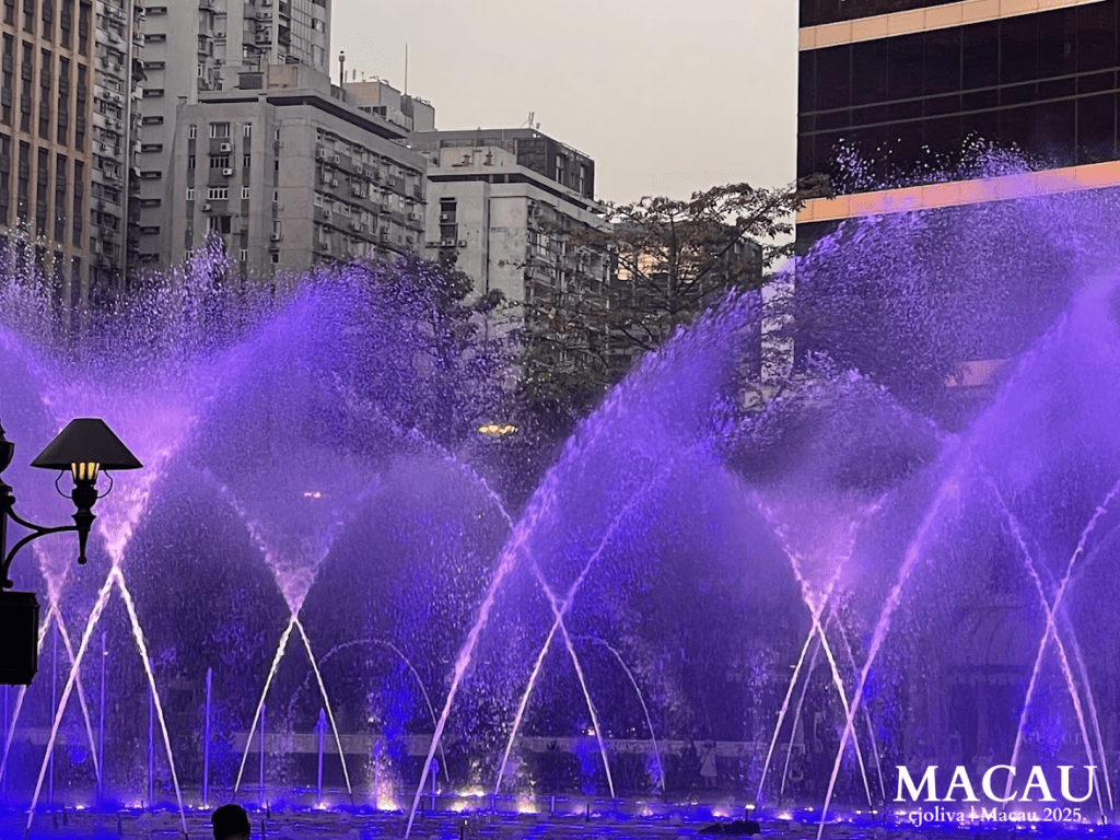 Illuminated fountain in Macau with purple and blue water jets arching across a city plaza at dusk, framed by high-rise buildings, trees, and a decorative street lamp.