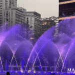 Illuminated fountain in Macau with purple and blue water jets arching across a city plaza at dusk, framed by high-rise buildings, trees, and a decorative street lamp.