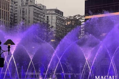Illuminated fountain in Macau with purple and blue water jets arching across a city plaza at dusk, framed by high-rise buildings, trees, and a decorative street lamp.